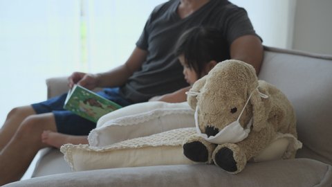 Plush Dog in Medical Mask on Sofa. Asian Little Girl and Father are reading a Picture Book. Stay Home. Covid-19 coronavirus pandemic, prevention epidemic