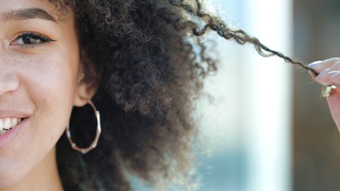 Close-up half face beautiful African girl afro curly hairstyle freckles brown eyes. Young ethnic American woman winding curl on finger, toothy smiling. Combing Сare for Afro Pigtails Brittle Hair