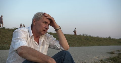 An elderly man sits on the lawn in the park against the background of walking people and looks thoughtfully into the distance, he touches his head with his hands and is sad alone among the people.