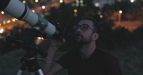 Astronomer with a telescope watching at the night sky with blurred city lights in the background.	