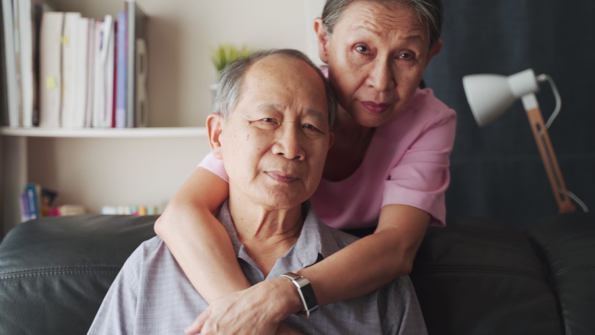 Portrait of Asian mature couple sitting and smiling in living room. Old woman hug man from behind and look at camera with happiness. Happy life after retirement, grandparents enjoy activity together. | Shutterstock HD Video #1056176609