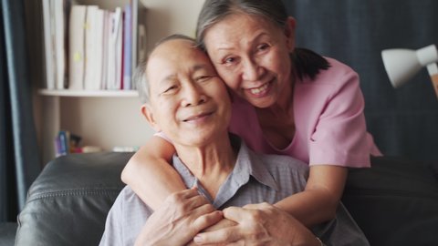 Portrait of Asian mature couple sitting and smiling in living room. Old woman hug man from behind and look at camera with happiness. Happy life after retirement, grandparents enjoy activity together.