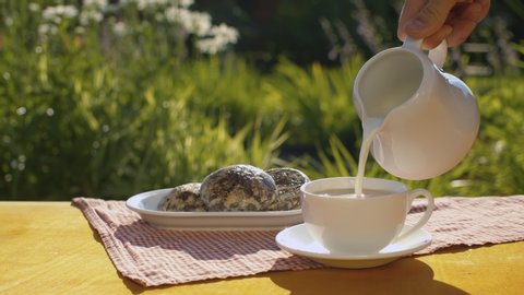 Adding milk into a cup of coffee. Close up hand holding a milk jug. Breakfast in the garden. The camera slowly moving on the slider