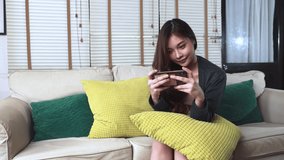 Young asian woman play game on her mobile. She is sitting on a sofa with yellow and green pillow around her.