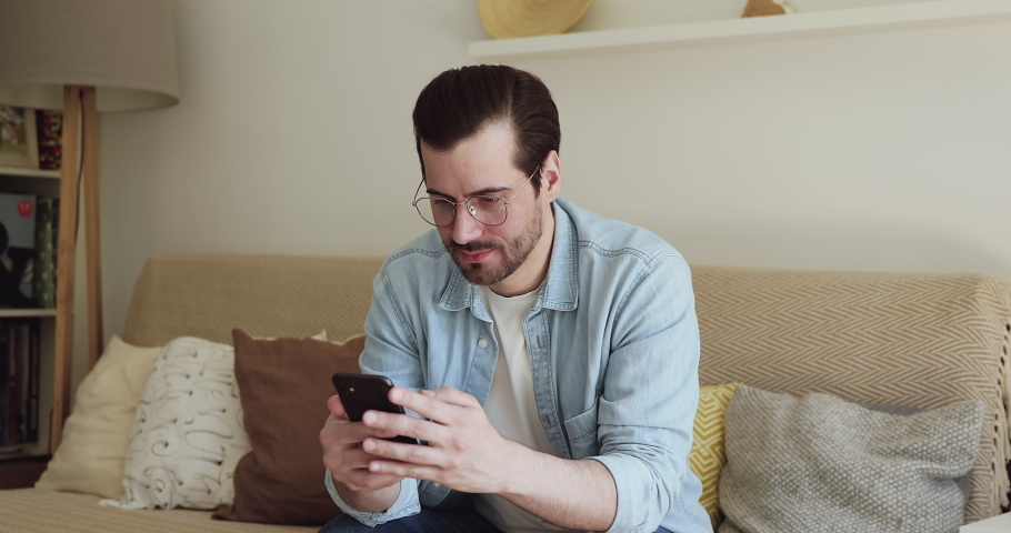 Young man wear glasses sit rest on couch in cozy living room holding mobile phone check messages read sms with super great news feels excited. Moment of victory, personal achievements, success concept Royalty-Free Stock Footage #1056178955