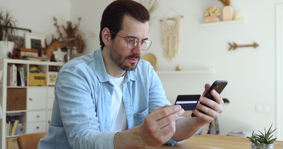 Millennial man in glasses sit at table hold smartphone and credit card frowns try to make payment through internet feels angry due lack of money, insufficient not enough funds. Victim of fraud concept Royalty-Free Stock Footage #1056178973