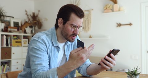 Millennial man in glasses sit at table hold smartphone and credit card frowns try to make payment through internet feels angry due lack of money, insufficient not enough funds. Victim of fraud concept