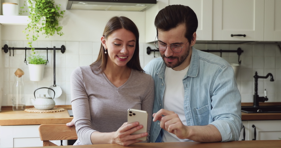 Couple sit at dining table in kitchen check lottery on-line app on smartphone see monetary win feels excited celebrating moment of victory. Sport game fan or online auction gambling winners concept Royalty-Free Stock Footage #1056178997
