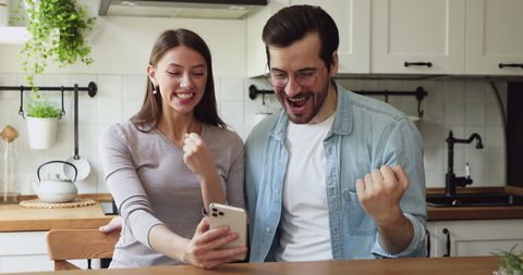 Couple sit at dining table in kitchen check lottery on-line app on smartphone see monetary win feels excited celebrating moment of victory. Sport game fan or online auction gambling winners concept