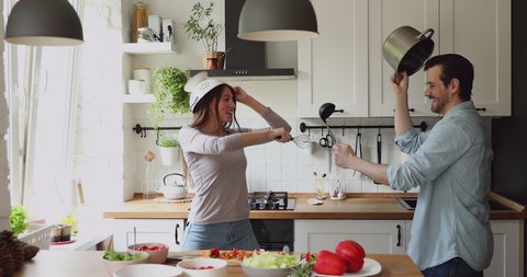 In modern home kitchen cheerful millennial couple take break from food preparation wear cookware holding kitchenware supplies fighting having fun laughing. Funny routine, happy family, cookery concept
