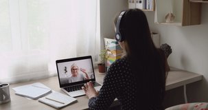 Rear view girl sit at desk wear headphones talk with aged female by video call, pc screen old granny enjoy distant conversation with granddaughter. Student and 50s tutor start class by videoconference