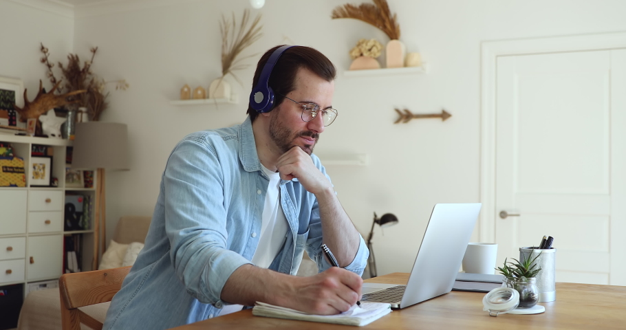 Millennial man sit at desk in living room looks at laptop screen listen audio through headphones use exercise book makes notes learn language develop improve gain new knowledge. Self-education concept Royalty-Free Stock Footage #1056179255