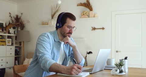 Millennial man sit at desk in living room looks at laptop screen listen audio through headphones use exercise book makes notes learn language develop improve gain new knowledge. Self-education concept