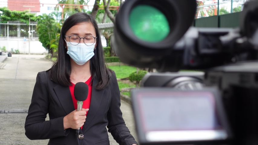 Reporter woman holding a microphone with reporting news and cameraman shooting outdoor news update while wearing  mask prevent Covid-19 or coronavirus quarantine pandemic. | Shutterstock HD Video #1056180407