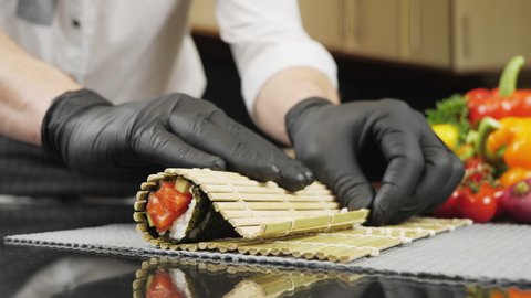 Japanese chef in black gloves at work preparing delicious sushi roll with salmon and avocado. Cook hands making Japanese sushi roll with bamboo mat. Delicious Japanese food.
