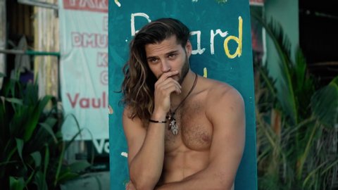 Young handsom European guy brunette with long hair with a wrapped sports body looks forward smiles with an embarrassed look and covers his face with his hand from shame