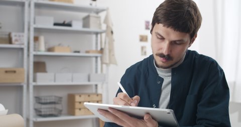Close up view of graphic designer holding notepad and drawing with stylus on it. Man with moustache holding digital tablet while creating illustration and sitting at table.