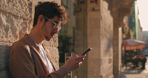 Good looking male tourist in glasses using mobile phone while standing leaned to wall. Crop view of millennial man with nose ring smiling while scrolling and typing screen at city street.