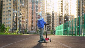 Slow motion video happy smiling caucasian little boy rides children's scooter, looks back at camera along sports road for running in Sunny summer weather against background of tall residential buildings