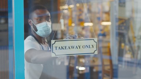 African coffee shop owner in medical mask and gloves hanging take away only sign on front door. Young afro entrepreneur opening cafe only for take away orders. Small business during quarantine concept
