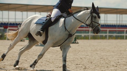 Young woman riding brown horse at show jumping competition on sandy parkour arena. Competitive rider galloping at training arena outdoor. Equestrian sport, slow motion.