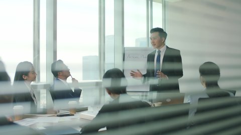 through the glass and blinds shot of a team of asian business people men and women meeting in modern office conference room
