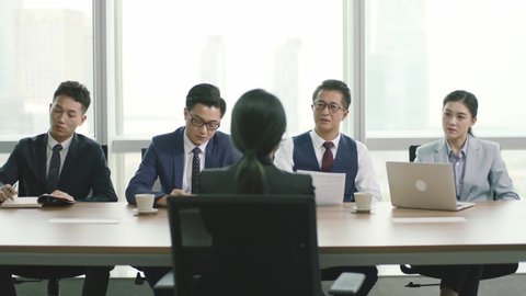 rear view of a young asian business woman being interviewed by a group of human resources executives in conference room of modern corporation