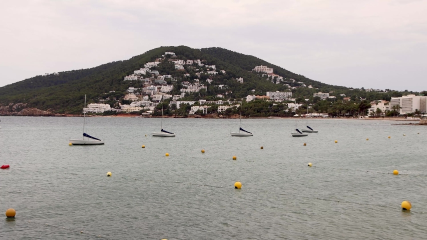 Footage of the coastal area of the town of Ibiza in Spain in the town of Santa Eularia des Riu showing a scenic view of hotels and boats in the ocean on a cloudy day