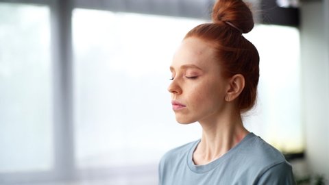 Close-up face of focused redhead young woman doing breathing yogic practices at the home office. closed eyes meditating on background of large window. Cute lady is making deep breath-exhalation.