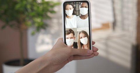 Happy friends take off medical mask during virtual meeting. Close up woman's hand holding smart phone in vertical position. Group video call screen collage. Remote communication online chat conference