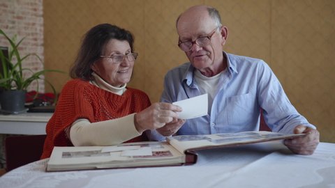 successful marriage, older husband and wife who retained love and care for each other are inspired by memory of years lived by reviewing photos in family photo album sitting at table in apartment