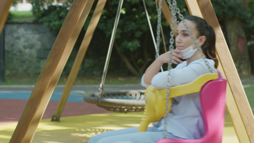 Thoughtful young girl child in the playground. She is swinging on a swing. She wears a medical mask to protect it from the virus.