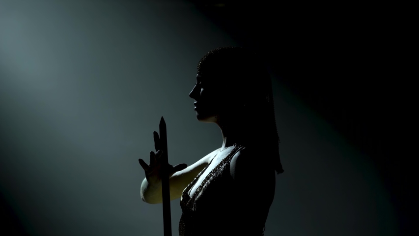 Silhouette of dancing Greek Goddess Artemis on stage in a dark studio with smoke and neon lighting. A bright young woman with a sword in a gold dress and a headdress. Dynamic neon lighting effects