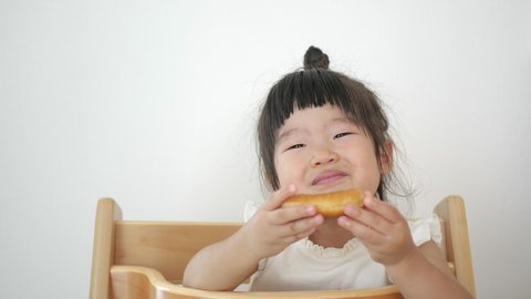Asian healthy preschool kid girl  in top knot hair eats donuts and smile. Sitting child chair. Happy toddler eating healthy organic and homemade food at home. Childhood, health concept