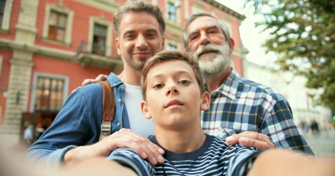 POV of cute teen Caucasian boy taking selfie photo with father and senior grandfather at street in town. Old man with beard with adult son and small grandson posing while making photos. Outdoor.