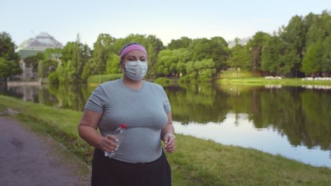 Overweight young woman in a medical mask running in park. Plus size female jogging outdoors in protective mask holding bottle of water. Weight loss during quarantine concept
