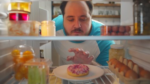 View from fridge of stout young man looking at donut but choosing fresh vegetables in container. Fat man on diet taking vegetables for snack. Will power, healthy diet and weight loss concept.