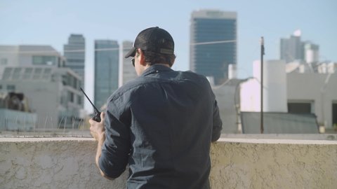Special agent security guard watching and reporting from a city rooftop with walkie talkie radio communication. Undercover man standing on top of a building during a mission or government surveillance