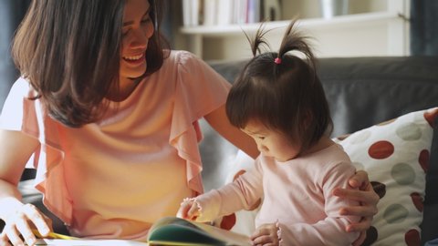 Asian beautiful young mother and her little daughter reading documentary book on sofa at home together. They pointing on the book looking it with happiness spending time together. Happy family concept
