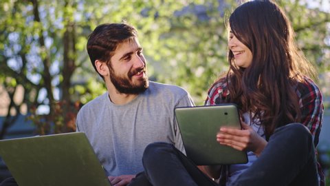 Attractive young female and good looking guy smiling large they chatting together while sitting on the grass outside at fresh air and working online from home using the tablet and laptop