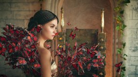 Beautiful divine majestic mystical young woman elf. Girl princess stands, backdrop medieval brick wall ancient castle. Creative elegant art vintage dress decorated with many artificial red butterflies