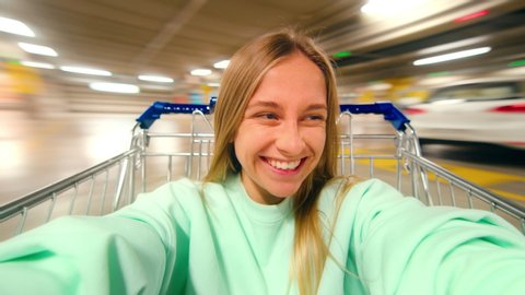 Excited happy young woman laugh and smile, sit inside shopping cart at parking lot of shopping mall or center. Generation z millennial teenager have fun on weekend, record selfie for social media