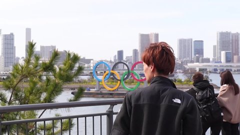 Tokyo, Japan - March 20, 2020 / People standing and walking by the esplanade  in Tokyo Bay where the famous Olympic rings sign is displayed. 
