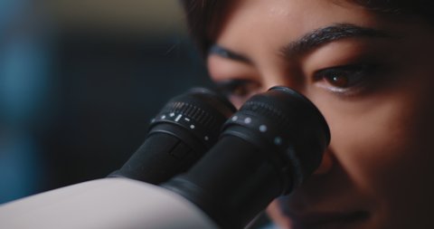 Close up of a female research scientist eyes, looking at samples under microscope.Blue lighting in a dark lab room.Slow motion, slider, shot with BMPCC 4K.Biochemistry, pharmaceutical medicine,science