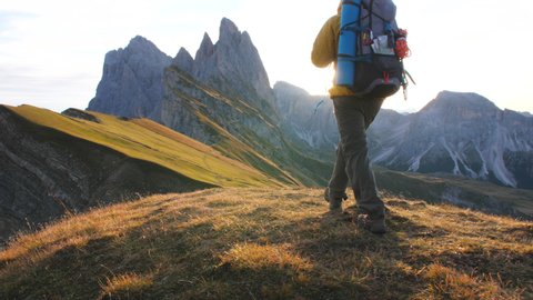 Young man hiking at Seceda mountain arriving to peak at sunrise. Backpack, yellow jacket, boots, beanie. Traveling to puez Odle, Dolomites, Trentino, Italy. Arkistovideo