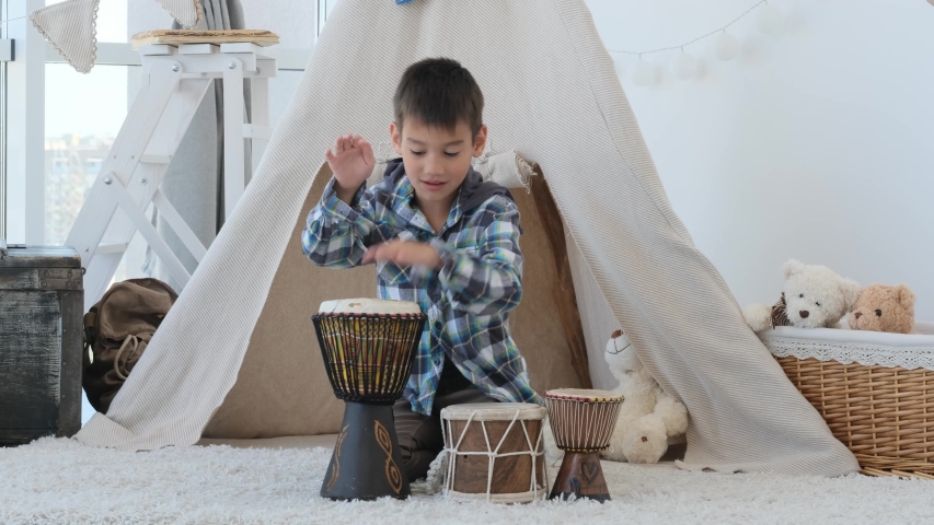 Little boy playing on ethnic drums djembe in room with wigwam