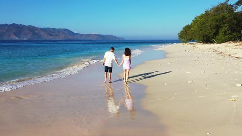 Young romantic couple holding hands while walking along the tropical white sand beach. Mountains in the background.