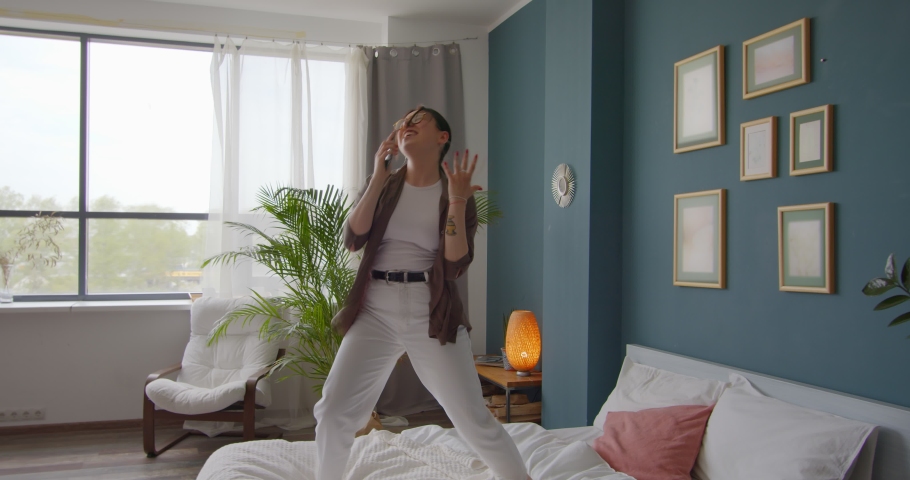 Happy woman dancing jumping on bed at home, having fun celebrating with funny dance. Singing karaoke on the phone and falling on the bed.  Royalty-Free Stock Footage #1056246707
