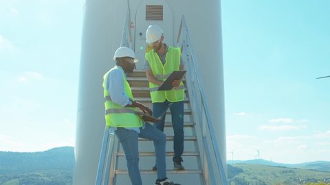 Two engineers men in uniform discuss use laptop stand near wind turbines ecological energy industry power windmill field worker renewable slow motion