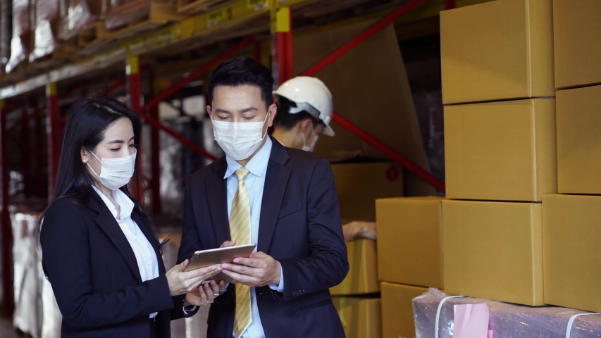 Asian business man and woman in suit and protective face mask talk and look at tablet with background of worker check stock in warehouse. Concept of new normal work after Covid 19 pandemic outbreak Royalty-Free Stock Footage #1056252833